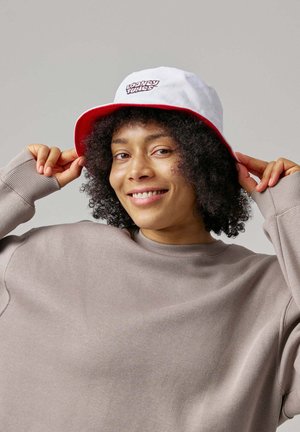 Person lächelt, während sie einen weißen und roten Looney Tunes Eimerhut anpasst und einen übergroßen beigen Pullover vor schlichtem Hintergrund trägt.