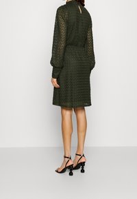 Tmavě zelené šaty vyrobené z texturované krajky, s dlouhými rukávy a staženým pasem. Černé sandály na pásky s kulatými, texturovanými podpatky.