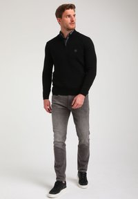 Schwarzer Zip-Kragen-Pullover mit einem strukturierten Rautenmuster, kombiniert mit grauen Slim-Fit-Jeans und schwarzen Sneakers mit weißen Sohlen.