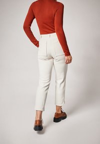 Pantalon blanc texturé taille haute avec deux poches arrière, associé à un haut ajusté à manches longues couleur rouille et des chaussures marron à lacets.