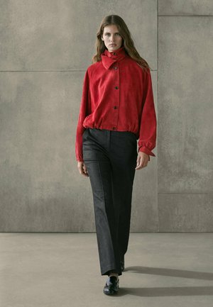 Donna che cammina con pantaloni neri, scarpe nere e una giacca rossa con bottoni e colletto alto, sullo sfondo di un muro di cemento grigio.