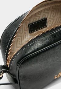 Zwarte leren handtas met een gestructureerd oppervlak, gouden rits en een lichtgekleurde binnenkant met een subtiel geometrisch patroon. Logo-tag zichtbaar.