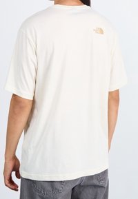 Krämfärgad bomulls t-shirt med korta ärmar, med en liten orange logotyp av "The North Face" på den övre ryggen. Enkel design, avslappnad passform.