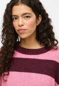 Strickpullover mit breiten rosa und dunkelbordeauxfarbenen Streifen, Rundhalsausschnitt und strukturiertem Stoff. Haare in lockeren, natürlichen Locken gestylt.