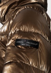 Manga de una chaqueta acolchada de bronce metálico con una etiqueta negra cosida que dice "Frieda & Freddies New York".