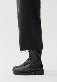 Zwarte leren veterschoenen met een stevige zool, voorzien van zichtbare stiksels, veterogen en een gladde afwerking. Gecombineerd met versleten zwarte jeans.