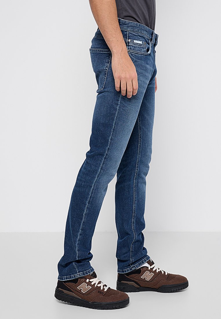 Schmal geschnittene blaue Denim-Jeans mit mittelhohem Bund und Fünf-Taschen-Design, das subtile Ausbleichungen und kontrastierende Nähte aufweist.