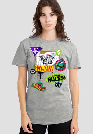 Woman wearing a gray Stranger Things T-shirt with colorful patches, including "Hawkins," "I make my own rules," and monster eye designs.