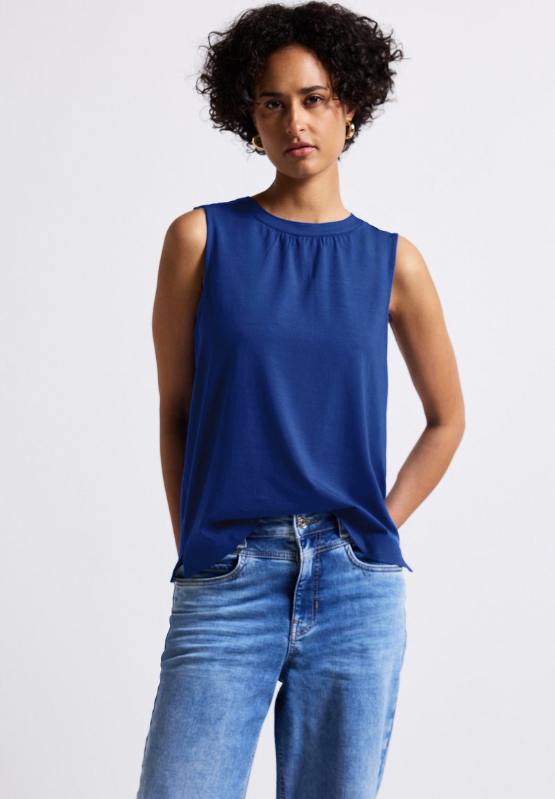 Top senza maniche di colore blu navy con scollatura arricciata, realizzato in un tessuto morbido. Abbinato a jeans in denim blu chiaro, con una vestibilità classica e cuciture visibili.
