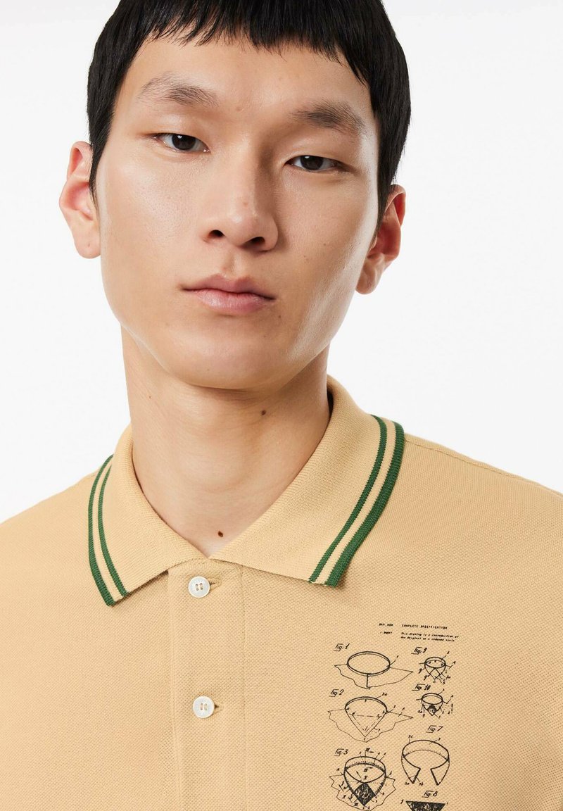 Camisa polo de tejido beige con un acabado texturizado, que cuenta con un cuello a rayas verdes y un estampado gráfico en el lado izquierdo.