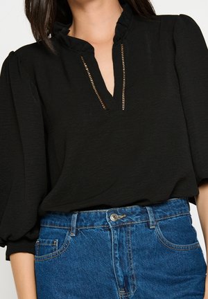 Kvinde iført en sort bluse med flæsekant i halsudskæringen og pufærmer kombineret med blå jeans med høj talje.