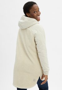 Lächende Person in einer cremefarbenen, gesteppte Kapuzenjacke mit Fleece-Rücken, die steht und über die Schulter schaut, vor einem einfarbigen Hintergrund.