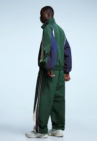 Vihreä verryttelytakki, jossa on tummansinisiä ja harmaita yksityiskohtia, korkeakauluksinen ja joustavat hihansuut, yhdistetty samansävyisiin, väljähköihin housuihin.