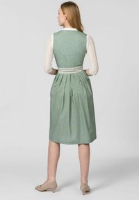 Grünes Kleid mit einem taillierten Oberteil, plissiertem Rock und Schärpe. Es hat einen gemusterten Stoff, lange Spitzeärmel und Schuhe in neutralen Tönen.