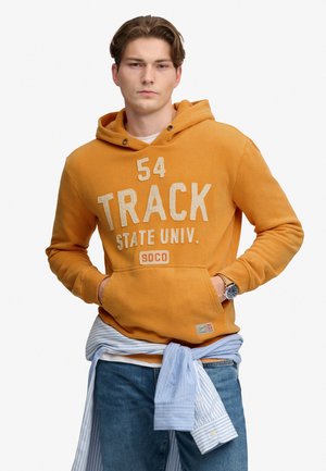Ung mand iført en sennepsgul hættetrøje med teksten "54 Track State Univ.", blå jeans og en lyseblå stribet skjorte bundet om taljen.