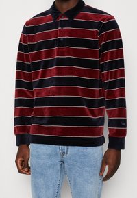 Man in een langemouwen, fluwelen polo met marineblauwe en bordeauxrode horizontale strepen, gecombineerd met lichtblauwe jeans.