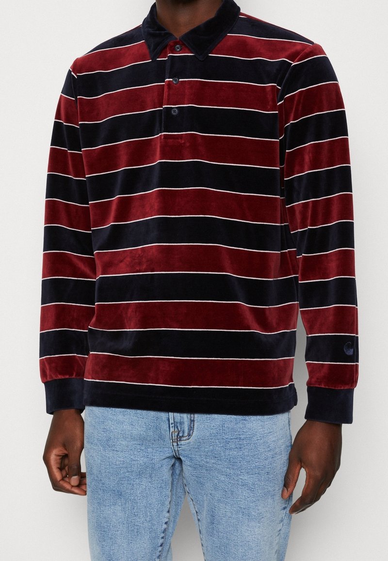 Man in een langemouwen, fluwelen polo met marineblauwe en bordeauxrode horizontale strepen, gecombineerd met lichtblauwe jeans.