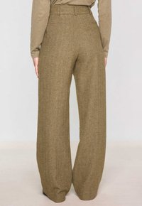 Vista posteriore di una persona che indossa pantaloni a gamba larga a vita alta in tessuto a spina di pesce marroni abbinati a una maglietta beige a maniche lunghe, in piedi contro uno sfondo semplice.