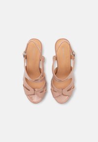 Nude lakované sandály na vysokém podpatku s otevřenou špičkou, které mají tři zakřivené výřezy a nastavitelný pásek kolem kotníku. Hladký povrch.