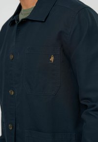 Chaqueta azul marino hecha de tela duradera con un bolsillo en el pecho que presenta un detalle bordado en oro y botones de metal en la parte frontal.
