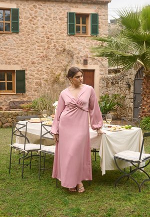 Kobieta w różowej sukience z długim rękawem stoi obok stołu jadalnego na zewnątrz, zastawionego jedzeniem i napojami, w ogrodzie z kamiennym domem i palmą w tle.