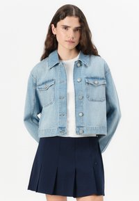 Lys blå denimjakke med en noe avkortet design, med to brystlommer, sølvknapper og spiss krage, båret over en hvit topp.