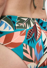 Κοντινή λήψη ενός ατόμου που φοράει πολύχρωμα μαγιό με σχέδιο τροπικών φύλλων σε αποχρώσεις μπλε, πράσινου, πορτοκαλί και ροζ.