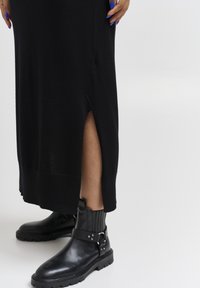 Vestito nero in maglia con fessura laterale, abbinato a stivali alla caviglia in pelle nera con dettagli in cinturino e fibbia. Texture liscia in generale.