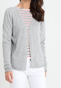 Cárdigan gris de punto con cuello en V y botones frontales, llevado sobre una camiseta de rayas rojas y blancas, combinado con pantalones blancos. Textura suave.