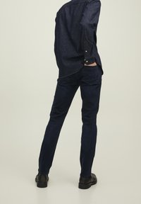 Person står med händerna i bakfickorna, iklädd mörkblå jeans, matchande denimskjorta och svarta skor på en neutral bakgrund.