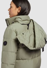 Geacă puffer în verde măsliniu deschis, cu guler înalt, închidere cu fermoar și capse, și un patch distinct pe mânecă. Material texturat.