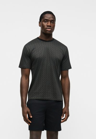 Homme portant un T-shirt ajusté gris foncé avec un petit texte répété et un short noir, debout devant un fond clair uni.
