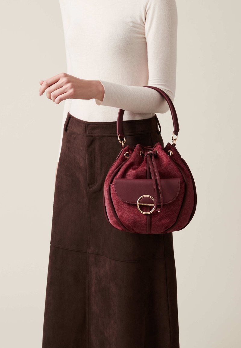 Borsa a mano bordeaux con silhouette arrotondata, materiale testurizzato, dettagli in hardware dorato, chiusura con coulisse e una piccola tasca frontale.