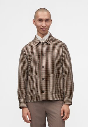 Man draagt een bruine en beige jas met hounds-tooth patroon en knopen over een witte blouse met kraag en lichtbruine broek, staande tegen een effen achtergrond.