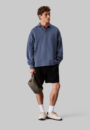Sudadera oversized de color azul claro con cuello, combinada con pantalones cortos negros. El modelo sostiene un bolso gris, lleva zapatillas beige y calcetines blancos.