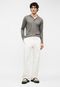 Jeune homme aux cheveux bouclés portant un polo gris à manches longues, un pantalon blanc et des mocassins beige-brun, debout avec une main dans la poche sur fond blanc.