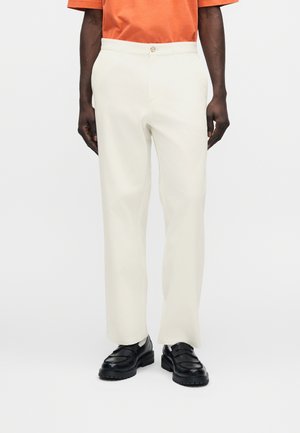 Mand iført cremefarvede bukser med lige ben, sorte loafers og en orange kortærmet skjorte, stående mod en hvid baggrund.