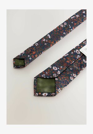 Florale Krawatte mit navy Hintergrund, verziert mit roten und weißen Blumen. Die Rückseite ist aus einfarbigem grünen Satin. Quadratische Enden und glatte Textur.