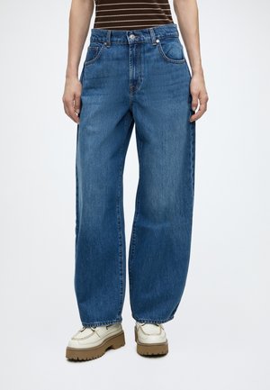 Person iført løse blå denimjeans, hvide platformssko med brune såler og en brun- og hvidstribet skjorte mod en hvid baggrund.