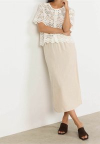 Žena v bílém krajkovém topu s krátkým rukávem na knoflíky, béžové midi sukni a černých sandálech s otevřenou špičkou a čtveratým podpatkem, stojící proti bílé zdi.