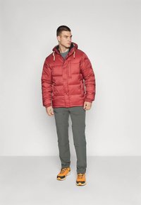 Sarkana pūdeļa jaka ar kapuci, ar teksturētu virsmu un aizdares akcentiem, kopā ar zaļām biksēm un oranžiem sporta apaviem.