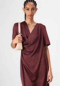 Draperet kjole med dyb V-hals i rig burgundy stof, korte ærmer og blød tekstur, parret med en beige taske med en slank rem.