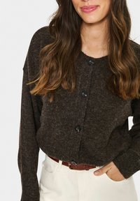 Frau mit langen braunen Haaren, die eine dunkelbraune, geknöpfte Strickjacke und weiße Hose mit einem braunen Gürtel trägt, die Hand in der Tasche.