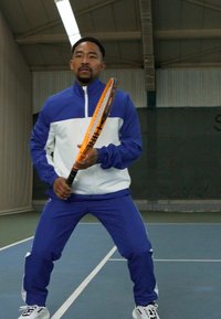 Vīrietis zilā un baltā sporta tērpā tur tenisista raketi, stāv uz laukuma ar teksturētu grīdu un iekštelpu apgaismojumu.