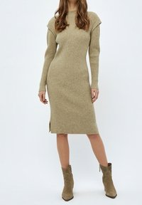 Robe tricotée en beige clair avec texture côtelée, silhouette ajustée, décolleté rond et fentes sur les côtés. Associée à des bottines beige à la cheville.