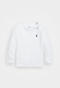 Vit långärmad skjorta i bomull med två silverknappar på vänster axel och en marinblå logotyp på vänster bröst.