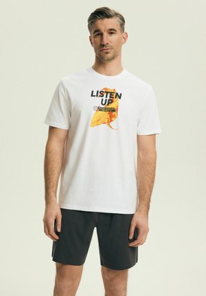 Mężczyzna stojący, ubrany w białą koszulkę z pomarańczową rybą i napisem "LISTEN UP" oraz czarne spodenki na jednolitym jasnym tle.