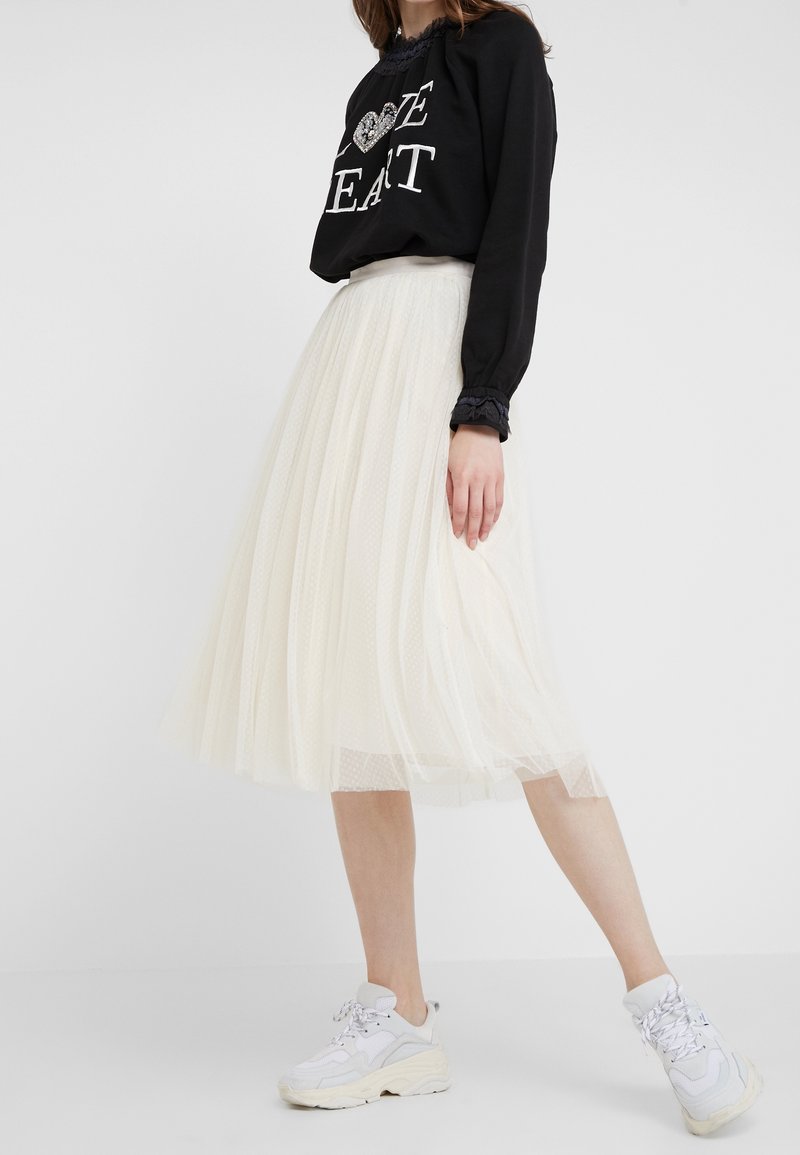 Sudadera negra con letras blancas y detalle de corazón, combinada con una falda de tul color crema hasta la rodilla y fluida, usada con zapatillas blancas gruesas.