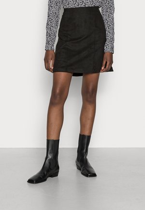 Gonna mini nera in suede con vita aderente, caratterizzata da cuciture verticali. Abbinata a stivali neri alla caviglia e a un top a maniche lunghe con fantasia.