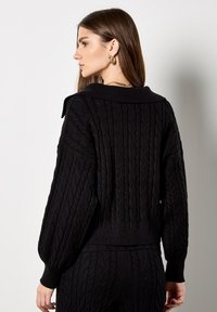 Jersey de punto negro con un amplio cuello, mangas abullonadas y dobladillo acanalado. La textura presenta patrones tejidos prominentes en toda su superficie.